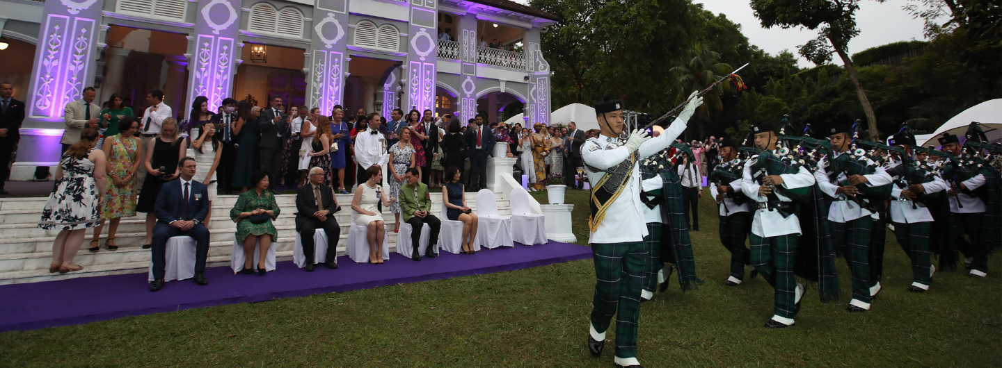 Bagpipe band marching on lawn, with seated and standing people in front of gray Victorian building.